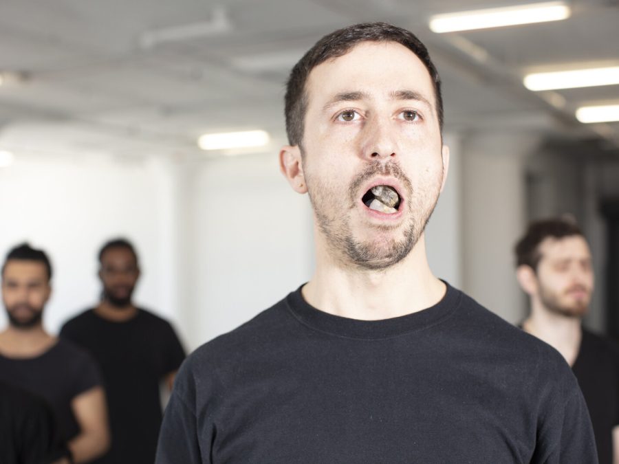 A group of people at varying distances from the camera are dressed in black shirts. The man in the foreground has his mouth open; inside are rocks.