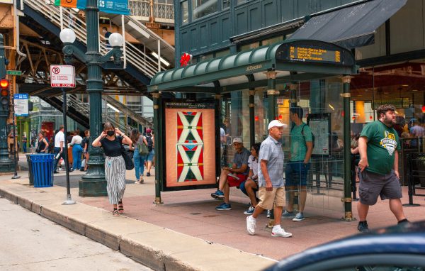 People walk past a bus shelter with an ad featuring a red, yellow, and black Native blanket
