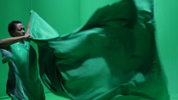 A person dressed in green in a green room holds a billowing green sheet in front of them