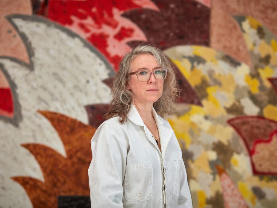 A light-skinned female-presenting person with wavy gray shoulder-length hair stands in front of a large painting with with wave-like shapes in pinks and whites