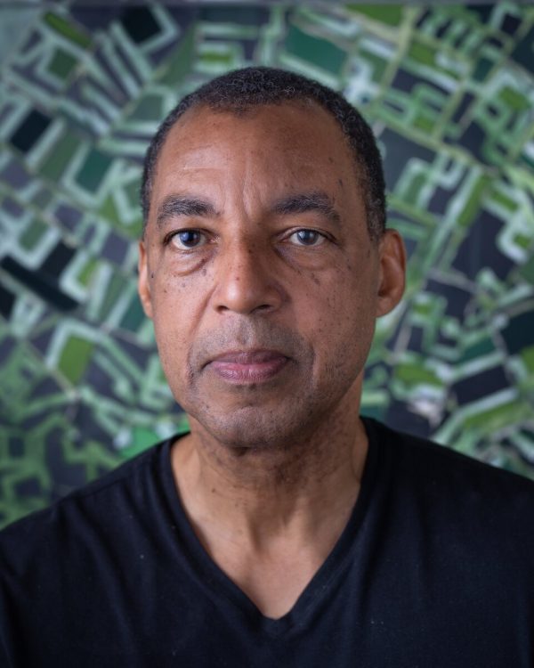 Shoulder length portrait of a male-presenting person with light brown skin and a shaved head posing in front of a green abstract artwork filled with white lines that seem to emanate from a central point behind him