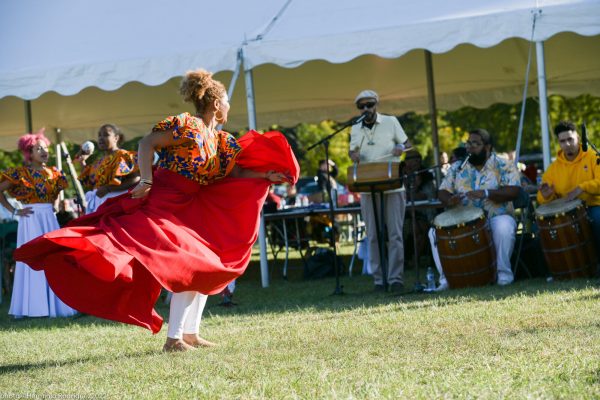A woman wearing a bright red flamenco-style dress dances on the grass in front of a group of people playing a variety of instruments.