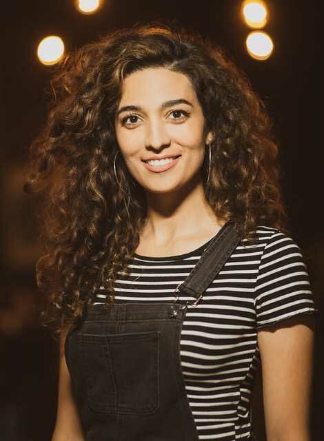 Night portrait of a female-presenting person with medium-light skin and full, curly brown hair wearing dark bibs and a black and white stripped shirt