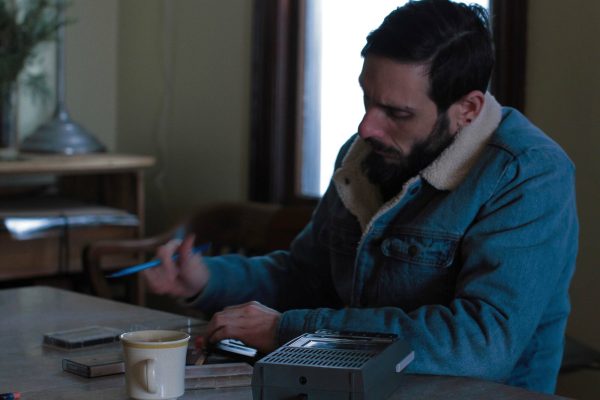 A brown-haired man sits at a table pen in hand. Despite being inside, he wears a coat.