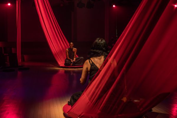 Two people sit on long red pieces of fabric facing each other in a red-lit room.