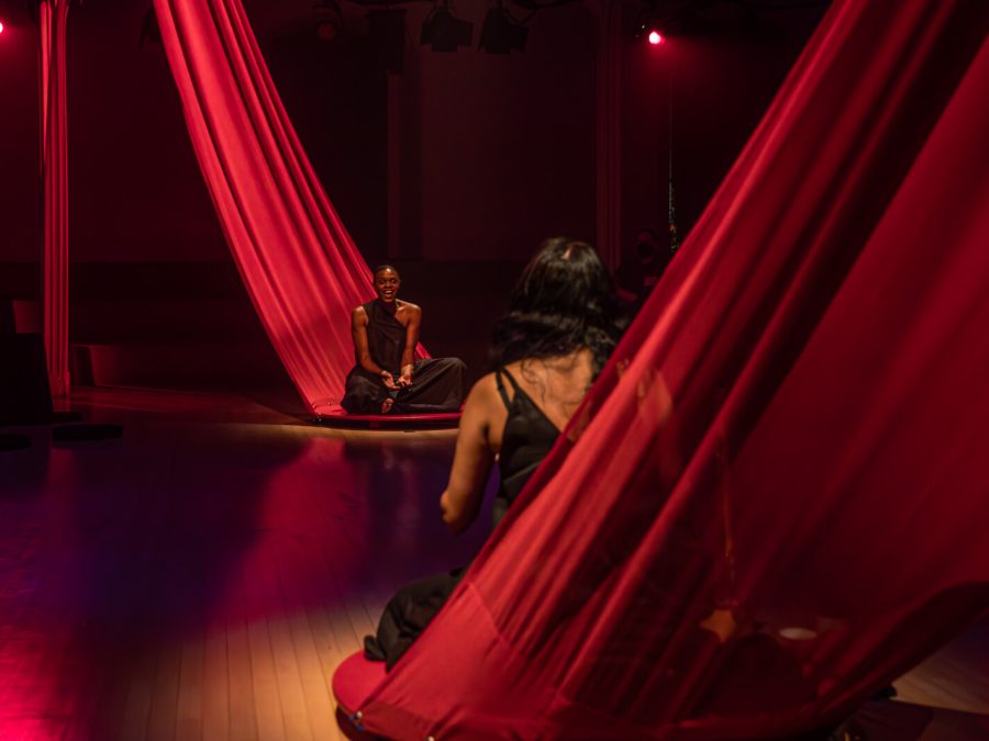 Two people sit on long red pieces of fabric facing each other in a red-lit room.