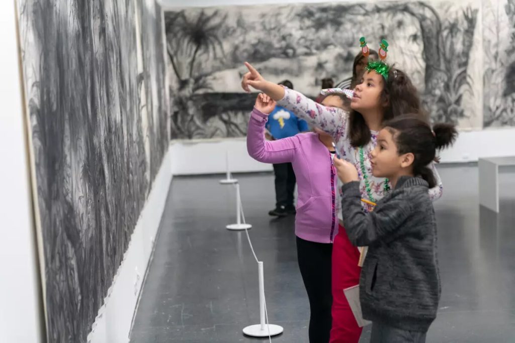 Children look and point at a black and white drawing of tropical trees and foliage that is displayed on two walls.