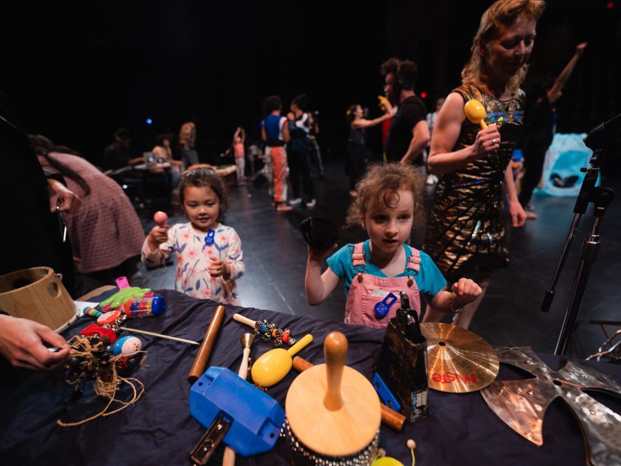 Kids and adults gather around tables filled with various noise making objects on a black stage.