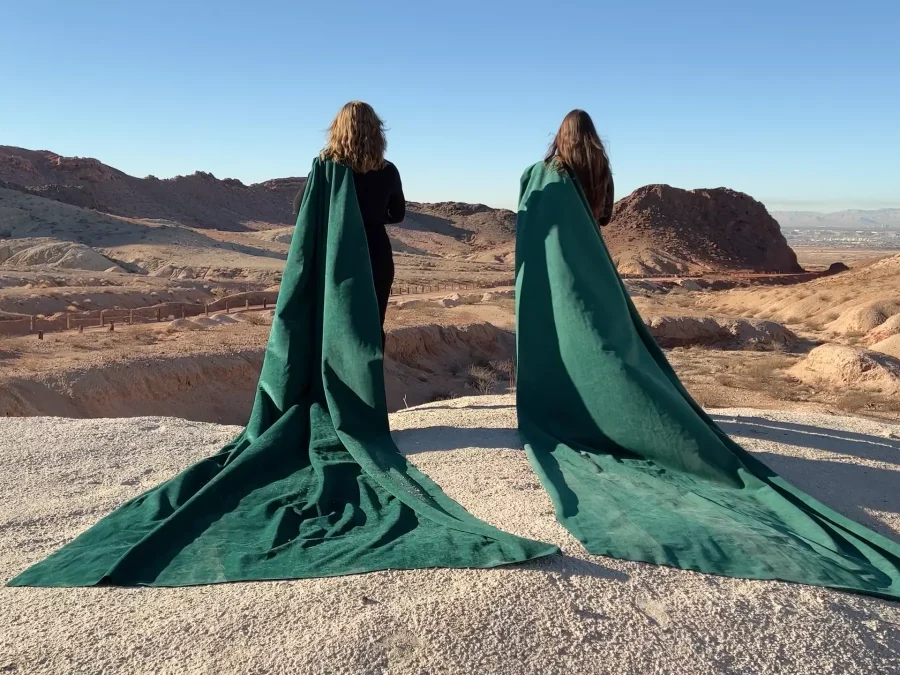 photograph of two people draped in long green capes facing a mountainous desert landscape