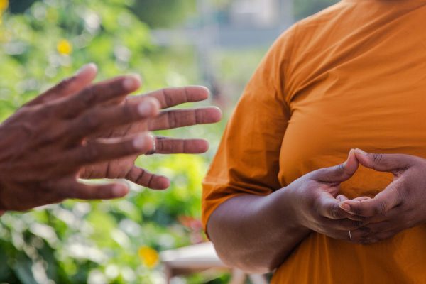 A pair of dark-skinned hands gestures toward another person in an orange shirt; only their torso can be seen, with their hands held a chest level in a bowl-like shape.
