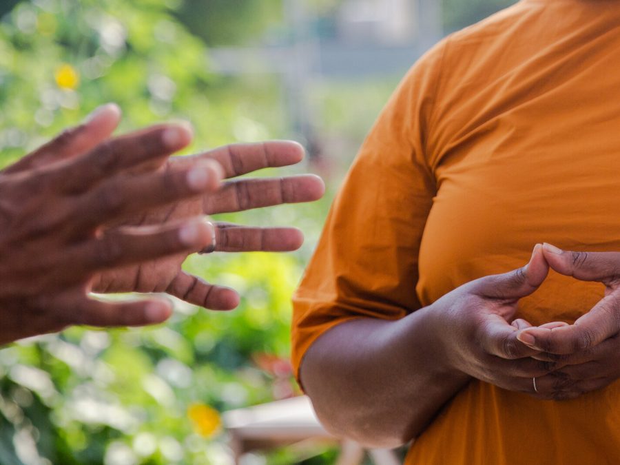 A pair of dark-skinned hands gestures toward another person in an orange shirt; only their torso can be seen, with their hands held a chest level in a bowl-like shape.