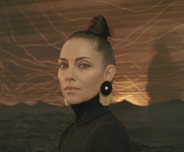 Teresita Fernandez, an olive-skinned woman with brown hair pulled into a tight bun a top her head, sits in front of a bronze and dark brown colored work in her studio in Brooklyn, NY. She wears a black turtleneck and dark, round earrings, both of which highlight her face.