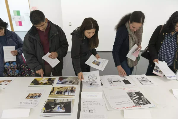 A line of people scan a table full of piles of paper. Some hold a few sheets in their hands.