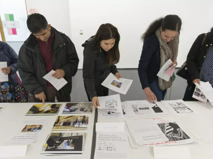 A line of people scan a table full of piles of paper. Some hold a few sheets in their hands.