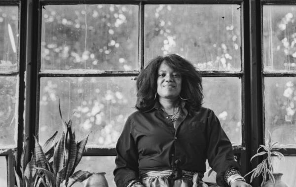 Black and white photo of a person with medium-length curly hair in front of large warehouse windows and surrounded by plants