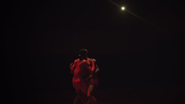 Two performers dressed in red in a dark room seeming to embrace.