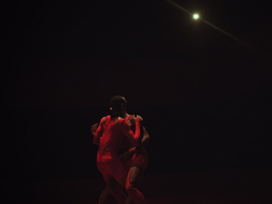 Two performers dressed in red in a dark room seeming to embrace.