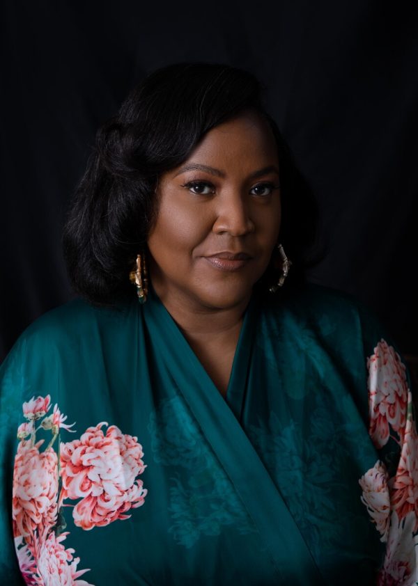 Image of a Black person with long black hair, silver earrings, and a silk flowery teal shirt.
