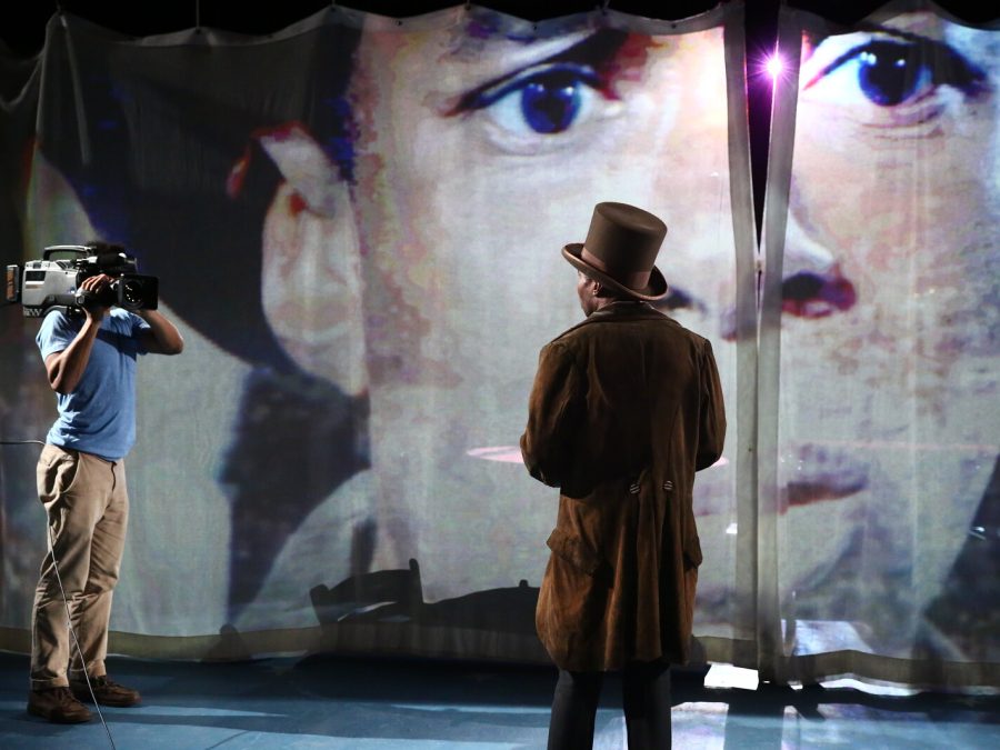 A person turned away from the camera in a top hat looks towards another person holding a video camera and a large screen displaying a man's head