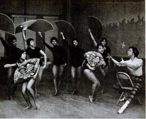 Archival photo of a group of people dancing; five people wearing black leotards stand in the back hold large dark fans in their right hand over their head