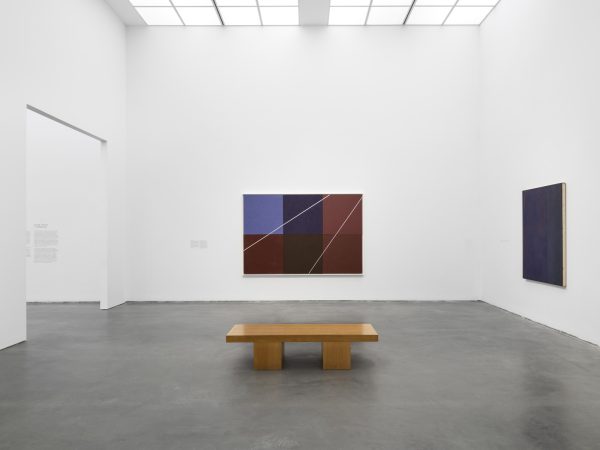 Gallery with two large paintings hung on adjoining walls and a wooden bench in the middle of the room
