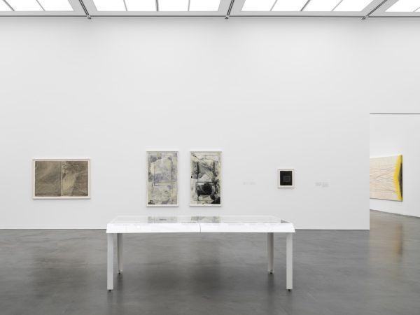 Gallery with four paintings hung on the wall and a display table in the middle of the room. A view through a doorway reveals another, larger painting.