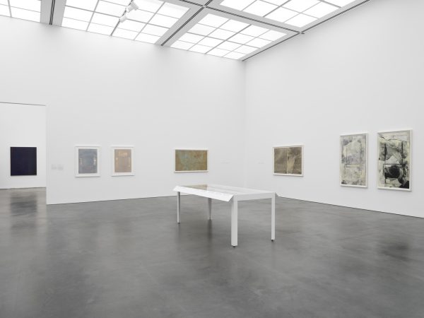 A gallery with several paintings hung on the walls and a display table in the middle. A view into another room reveals another painting.
