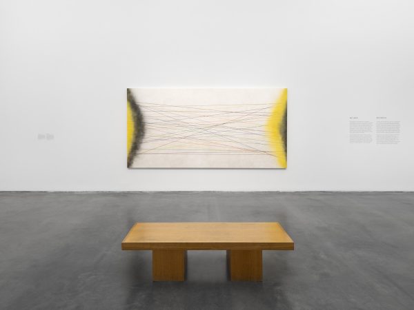 A gallery wall with a singular large painting hung on it. A wooden bench has been placed in front of the work.