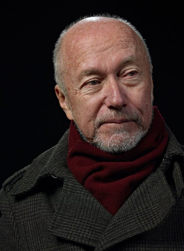 Portrait of a mostly bald man with a white goatee in a burgundy scarf and grey peacoat against a black background.