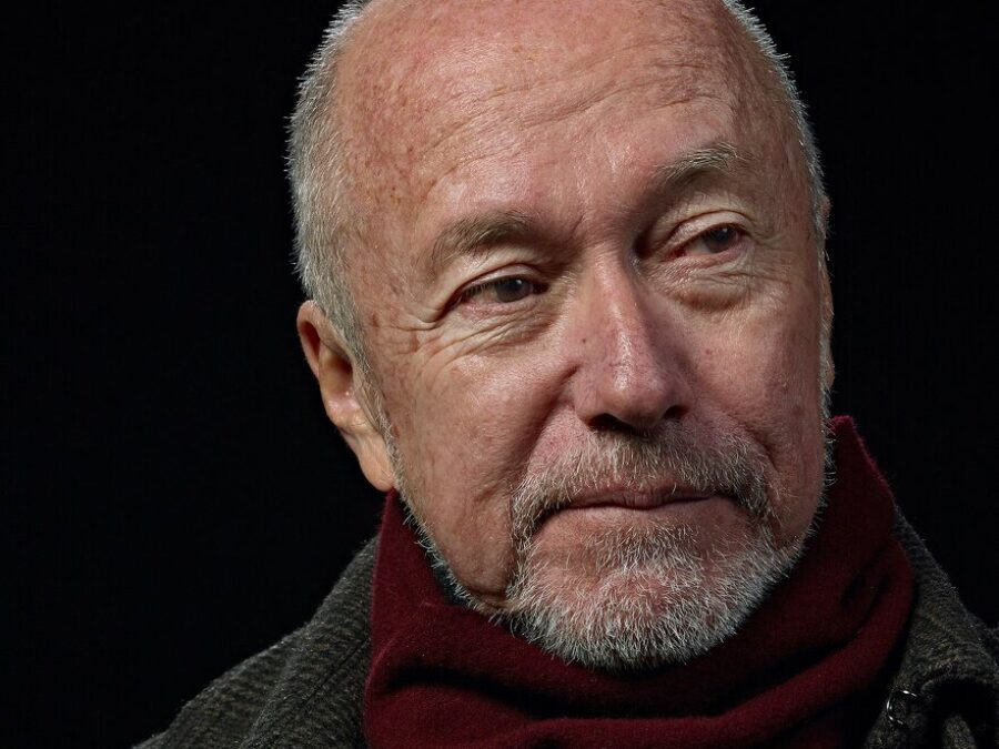Portrait of a mostly bald man with a white goatee in a burgundy scarf and grey peacoat against a black background.