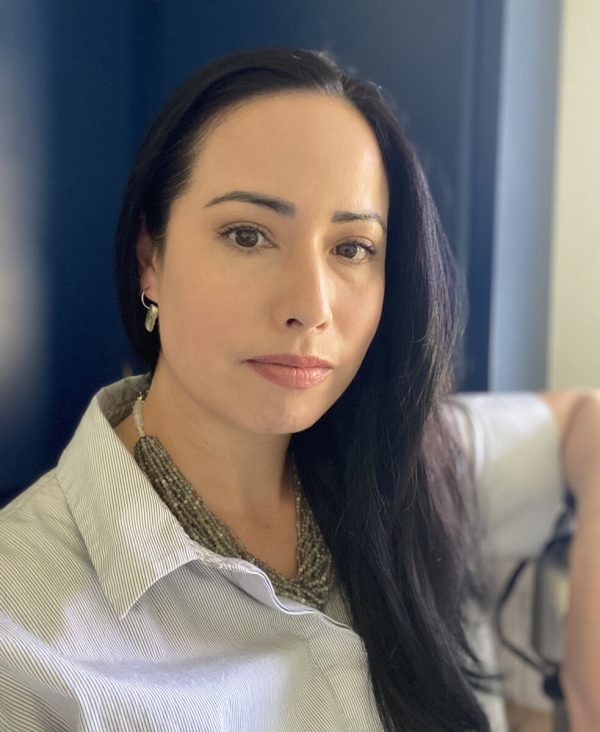 Portrait of a female-presenting person with long dark hair posing with their left arm resting on an object as tall as their shoulder