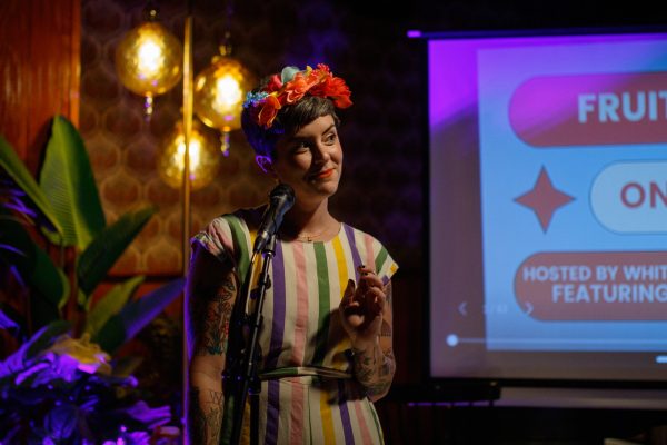 A woman in a white, green, yellow, pink, and purple dress with red flowers in her hair stands in front of a microphone stand. She looks off to her left. Behind her, a portion of a large projection screen appears to the right.