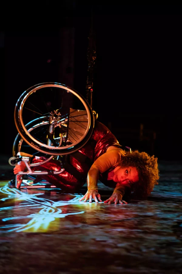 Alice Sheppard, a multiracial Black woman with coffee-colored skin and short curly hair, crouches onstage beneath her chair, her legs and the wheels curled toward her core, arms reaching toward the camera. She peers intently beyond the camera, eyebrows raised.