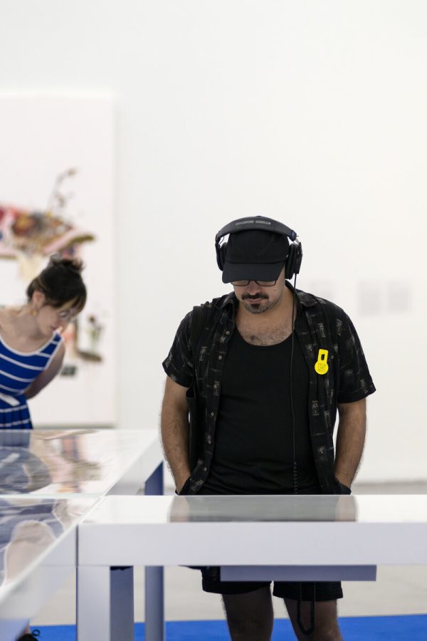 A man wearing headphones looks down at a display case within a white-walled gallery space.