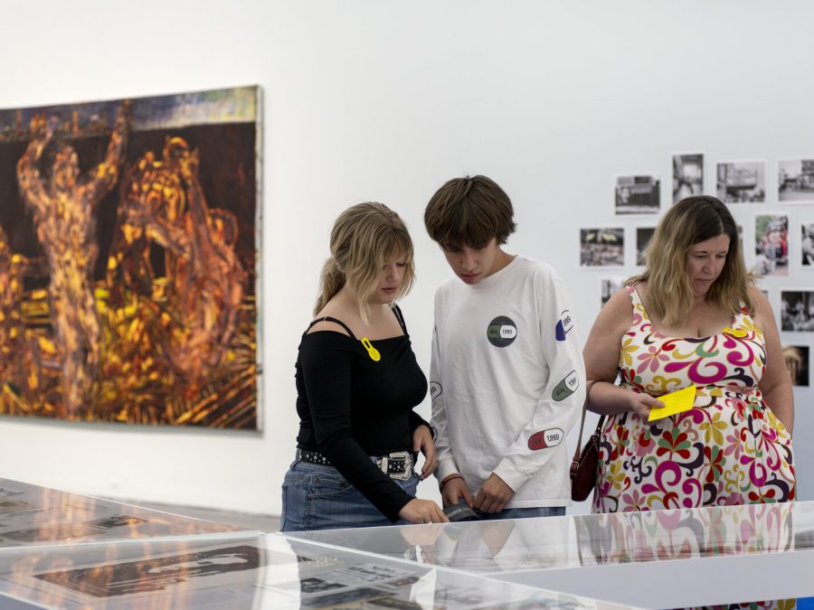 Three people look at a collection of object in display cases. Behind them, several artworks hang on a white wall.
