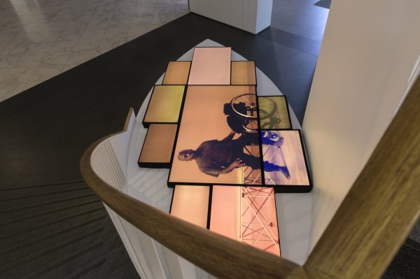 Sideways view of ten TV screens in a tight arrangement that together display a still image of a person next to a wheelchair.
