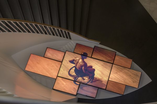 An array of TV screens at the foot of the MCA spiral staircase all show a Black man in a wheelchair whose back is to the viewer.