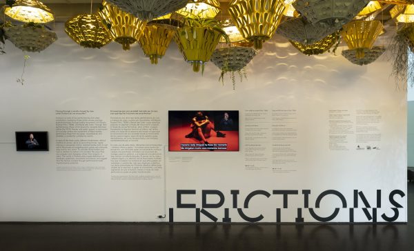 A wall with two videos and a lot of text underneath a ceiling filled with hanging baskets and lights.