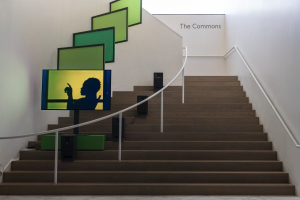 A series of screens of various greens ascends a staircase. The front screen features a shadow profile of a person sitting with one arm raised in a 90-degree angle.