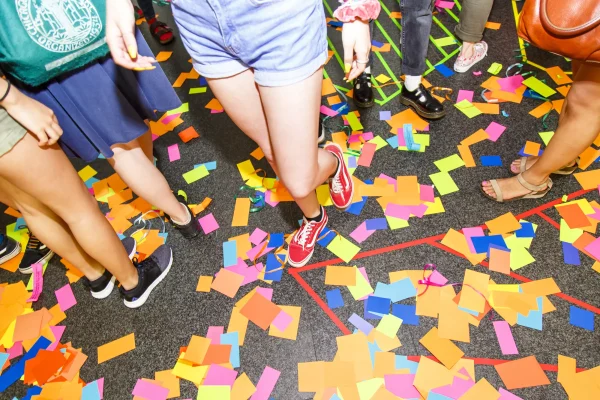 The bottom half of several people as they walk over multicolored scraps of paper.