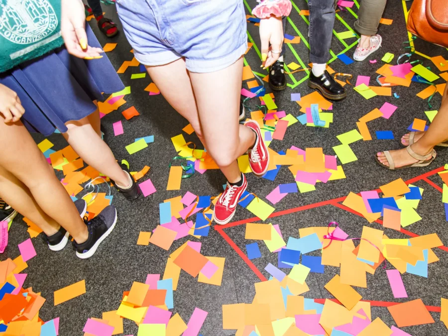 The bottom half of several people as they walk over multicolored scraps of paper.