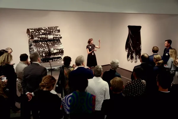 A group of people gather around someone speaking in a small gallery space