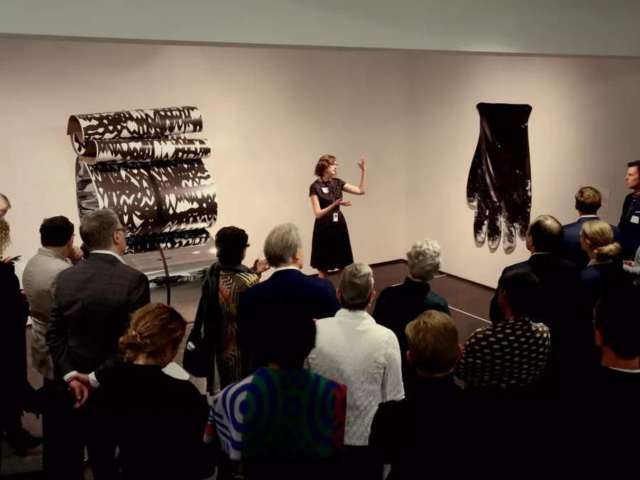 A group of people gather around someone speaking in a small gallery space