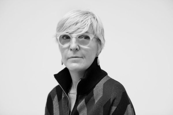 Black and white portrait of a female-presenting person with light skin, short light hair, and light glasses in a dark shirt with their collar popped