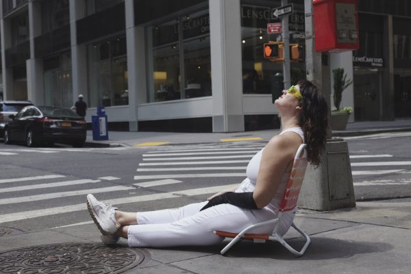 A woman with dark brown hair sits in a beach chair on the corner of an empty urban street, kitty corner from a building labeled Lenscrafters. She wears all white, minus a singular long, black glove and neon yellow sunglasses.