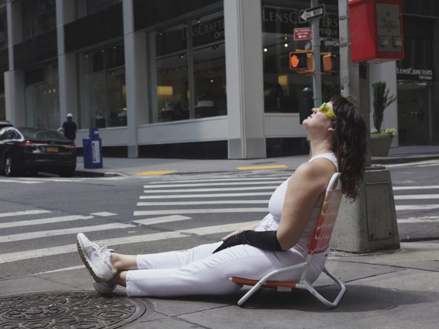 A woman with dark brown hair sits in a beach chair on the corner of an empty urban street, kitty corner from a building labeled Lenscrafters. She wears all white, minus a singular long, black glove and neon yellow sunglasses.