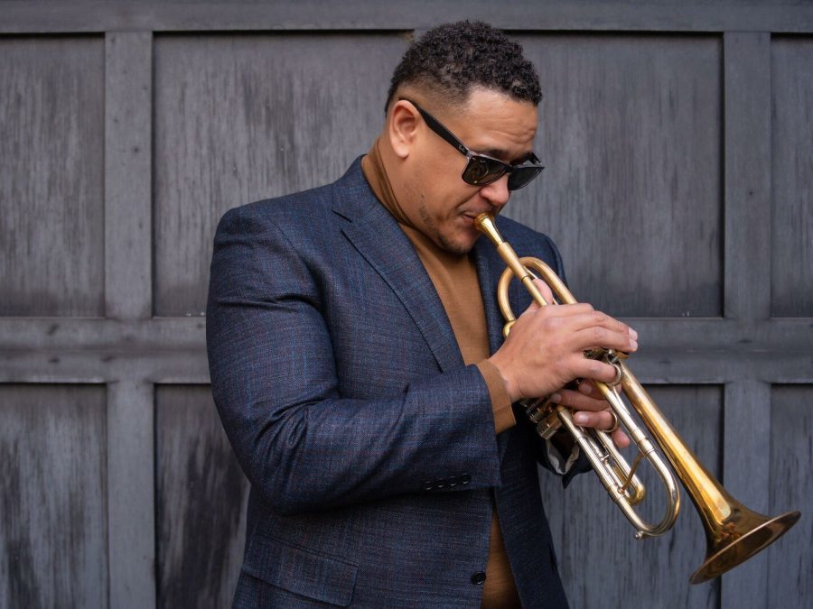 A Black man with dark glasses in a blue blazer blows into a trumpet.