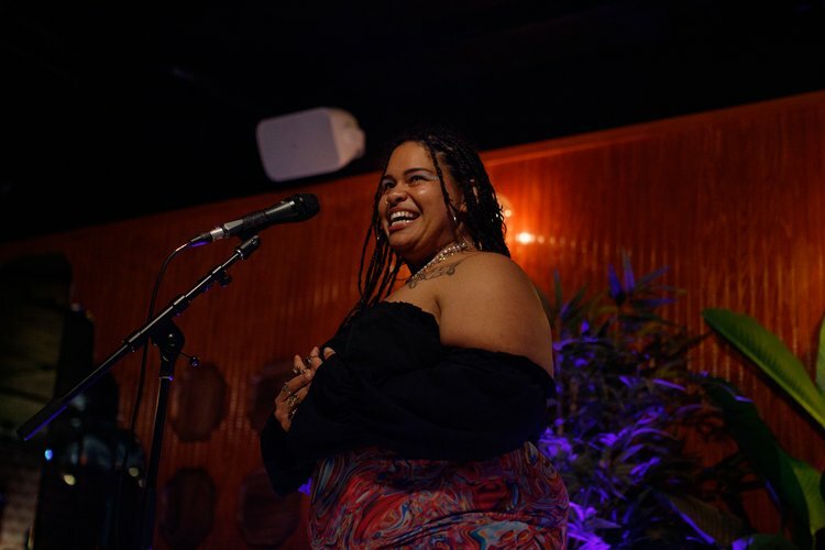 A person with long, dark braids smiles while standing in front of a microphone stand.