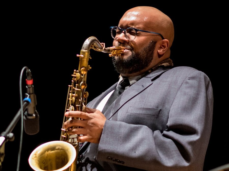 A bald, medium-skin toned man plays the saxophone; his face is scrunched up from blowing.