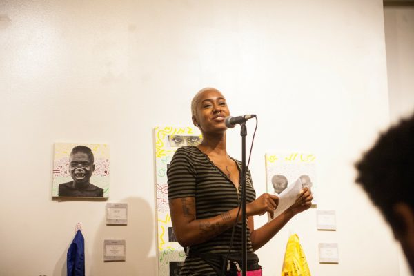 A brown-skinned person with short blond hair stands in front of a microphone with papers and images taped to a white wall behind them.